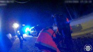 Policjant i ratownik medyczny podczas udzielania pomocy mężczyźnie leżącemu na noszach. W głębi pojazdy służb ratunkowych i ludzie.