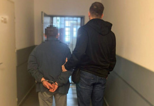 Funkcjonariusz prowadzi zakutego w kajdanki zatrzymanego mężczyznę korytarzem.