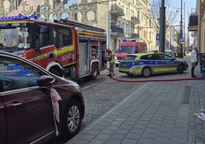 Radiowóz policyjny i dwa wozy strażackie stoją na ulicy przed budynkiem gdzie wybuchł pożar.