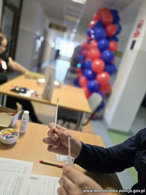 Kobiety siedzą za stołem , pobierany patyczkiem materiał genetyczny. Widać logo policji i fundacji DKMS