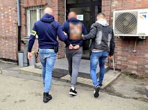 Dwaj funkcjonariusze prowadzą zatrzymanego mężczyznę zakutego w kajdanki do budynku z cegły.