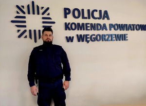 Policjant stoi na tle ściany z napisem Policja Komenda Powiatowa Policji w Węgorzewie.
