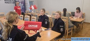 Na pierwszym planie trzy policjantki siedzące przy stole, z których jedna trzyma tabliczkę z napisem DKMS. Z tyłu widać stojącą kobietę i dwie inne osoby siedzące przy stole.
