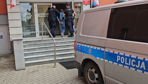 Dwaj policjanci prowadzą zatrzymaną osobę po schodach do budynku.