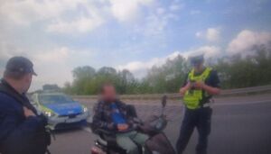Mężczyzna na elektrycznym wózku inwalidzkim rozmawia z dwoma policjantami. W tle na drodze stoi radiowóz policyjny.