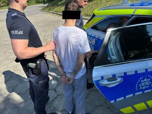 Zatrzymany wsiada do radiowozu. Z tyłu stoi policjant.
