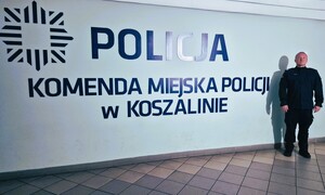 Policjant pozuje na ściance z napisem: Policja, Komenda Miejska Policji w Koszalinie.