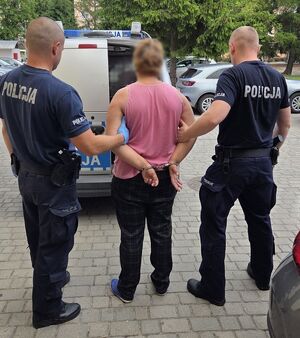 Dwaj policjanci prowadzą zatrzymaną. W tle widać policyjny radiowóz.