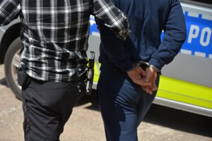 Policjant prowadzi zatrzymanego mężczyznę zakutego w kajdanki.