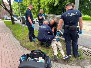 Policjanci udzielają pomocy poszkodowanemu w wypadku leżącemu na trawniku przy ulicy.