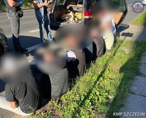 Mężczyźni siedzą na krawężniku, przed nimi stoją dwaj policjanci, a obok nich zaparkowane auto.