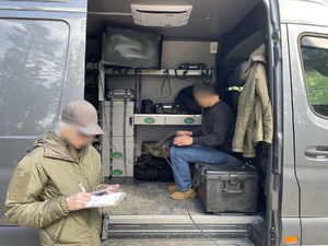 Policjant siedzi w busie i na kolanach trzyma laptopa, przed busem stoi inny funkcjonariusz.