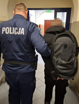 Policjant prowadzi korytarzem zatrzymanego mężczyznę zakutego w kajdanki.