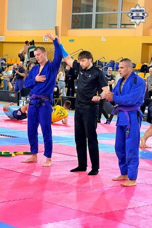 Dwaj sportowcy z sędzią na macie. Sędzia ogłasza zwycięzcę, podnosząc jego rękę do góry.