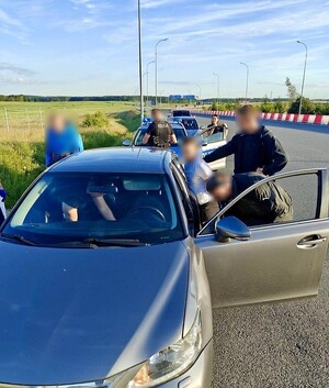 Policjanci z zatrzymanym przy samochodzie. W głębi na drodze radiowozy.