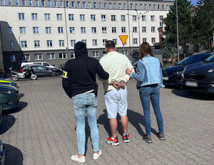 Policjanci prowadzą zatrzymaną osobę, która na rękach trzymanych z tyłu ma założone kajdanki.