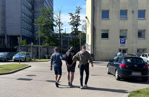 Policjanci prowadzą zatrzymaną osobę, która na rękach trzymanych z tyłu ma założone kajdanki.