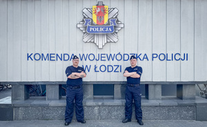 Dwaj policjanci pozują do zdjęcia przed obeliskiem z napisem Komenda Wojewódzka Policji w Łodzi.