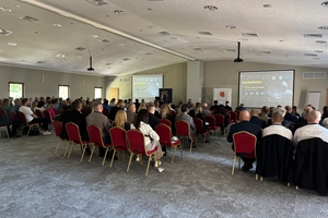 Uczestnicy konferencji siedzą na krzesłach zwróceni w kierunku ściany, na której wiszą ekrany wyświetlające prezentację. Przy mównicy stoi prelegent.