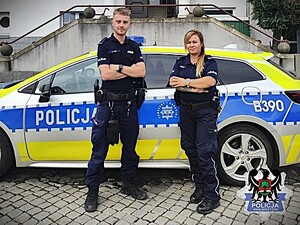 Policjantka i policjant stoją przed radiowozem i pozują do zdjęcia.