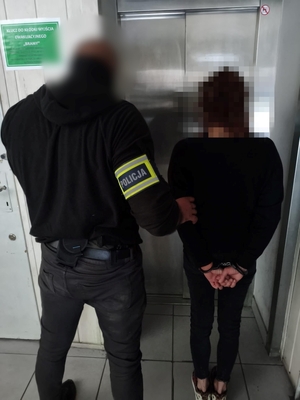 Policjant prowadzi zatrzymaną osobę zakutą w kajdanki.