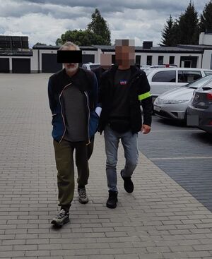 Zatrzymany mężczyzna prowadzony przez policjanta przez parking obok zaparkowanych samochodów.