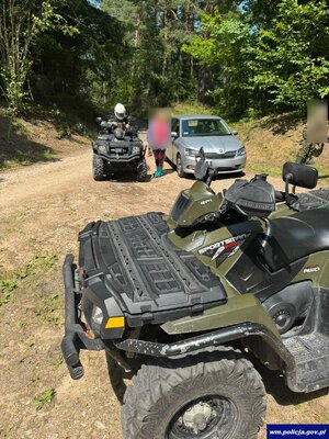 Policjant na quadzie, stojący obok mężczyzna i auto na leśnej drodze.