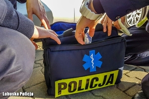 Policyjna torba medyczna z napisem Policja, na żółtym odblaskowym tle. Dwie osoby zaglądają do torby i wyciągają lub przeglądają wyposażenie medyczne.