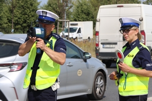 Stojący przy radiowozie policjant z ręcznym miernikiem prędkości i policjantka z tzw. lizakiem podczas kontroli drogowej.