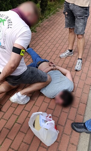 Zatrzymany leży na chodniku, obok stoją dwaj funkcjonariusze. Przy zatrzymanym leży reklamówka z zabezpieczonymi przez policjantów przedmiotami.