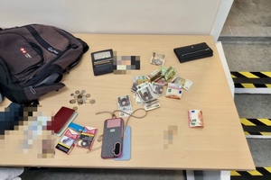 Na blacie leżą zabezpieczone przez policjantów przedmioty: banknoty, monety, dwie paczki papierosów, dwa smartfony, dwa portfele i plecak.