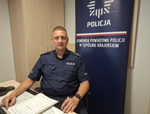 aspirant Zbigniew Pastwik siedzi przy biurku, na którym lezą dokumenty. W tle za nim przy ścianie baner z napisem Komenda Powiatowa policji w Sępólnie Krajeńskim.