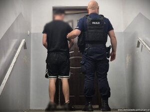 Policjant z zatrzymanym mężczyzną stoi przed drzwiami mieszkania na klatce schodowej.