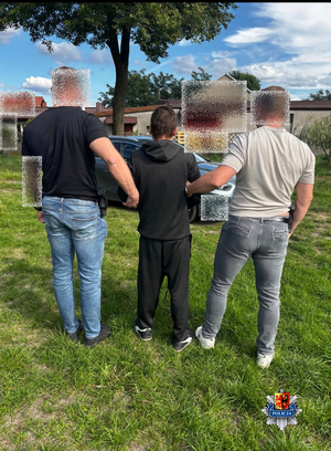 Dwaj funkcjonariusze trzymają zatrzymanego mężczyznę zakutego w kajdanki.