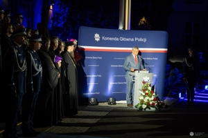 Uczestnicy uroczystości stoją na schodach wejściowych Komendy Głównej Policji obok przy mównicy przemawia sekretarz Stanu w Ministerstwie Spraw Wewnętrznych i Administracji Wiesław Szczepański.