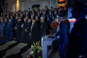 Komendant Główny Policji przemawia podczas uroczystości stojąc przy mównicy. w głębi Uczestnicy uroczystości stoją na schodach wejściowych do budynku Komendy Głównej Policji.