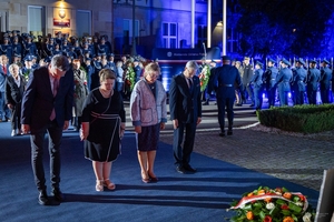 Cztery osoby oddają hołd poległym policjantom przed obeliskiem na terenie Komendy Głównej Policji. W tle uczestnicy uroczystości stojący przed budynkiem KGP.