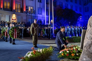Mężczyzna składa wieniec pod obeliskiem na terenie Komendy Głównej Policji. W tle uczestnicy uroczystości stojący przed budynkiem KGP.