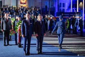 Delegacje z wieńcami przed obeliskiem na terenie Komendy Głównej Policji. W tle uczestnicy uroczystości stojący przed budynkiem KGP.