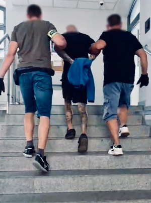 Policjanci wprowadzają po schodach do budynku zatrzymanego mężczyznę zakutego w kajdanki.