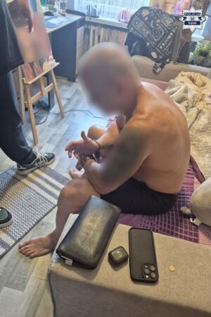 Zatrzymany mężczyzna w kajdankach n rękach siedzi na łóżku. Obok niego stoi policjant.