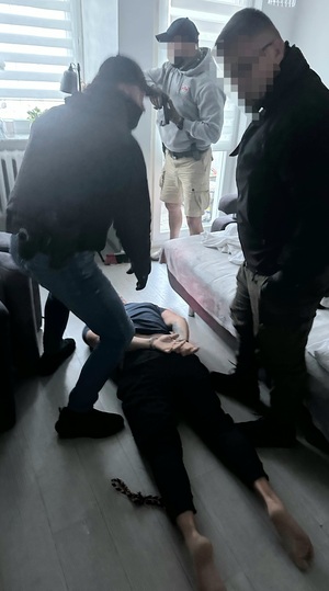 Zatrzymany mężczyzna leży na podłodze w mieszkaniu zakuty w kajdanki. Wokół niego stoją trzej funkcjonariusze.