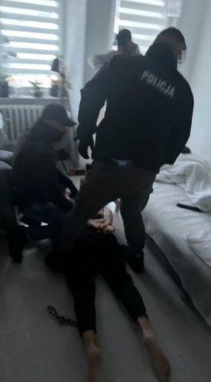 Zatrzymany mężczyzna leży na podłodze w mieszkaniu zakuty w kajdanki. Wokół niego stoją trzej funkcjonariusze.