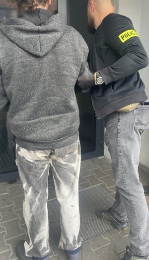 Policjant w prowadza do budynku zatrzymanego mężczyznę w kajdankach.