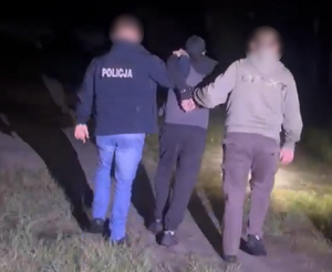 Dwaj funkcjonariusze prowadza zatrzymanego mężczyznę zakutego w kajdanki.