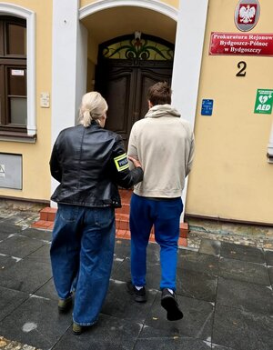 Funkcjonariuszka prowadzi zatrzymanego do budynku prokuratury.
