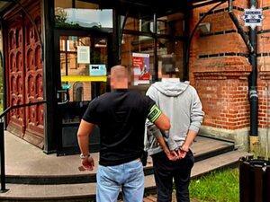 Nieumundurowany policjant prowadzi zatrzymanego w kierunku drzwi wejściowych do budynku.