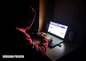 Mężczyzna w bluzie z kapturem przy laptopie w ciemnym pomieszczeniu.