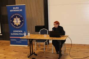 Kobieta siedzi przy stole, przed sobą ma otwarty laptop. Obok niej puste krzesło. Po lewej stronie banner z logiem Biura Kryminalnego Komendy Głównej Policji.