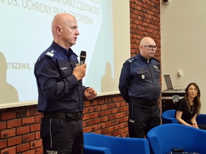 dwaj policjanci stoją na tle ekranu z prezentacją, kobieta siedzi w fotelu